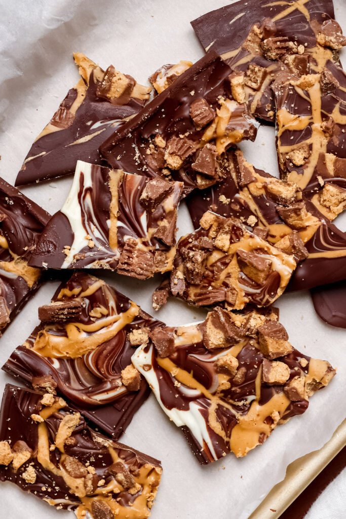 A batch of Reese’s Chocolate Bark broken into pieces, showing swirls of dark, semi-sweet, and white chocolate with a peanut butter drizzle and chopped Reese’s cups on top. A perfect no-bake dessert.