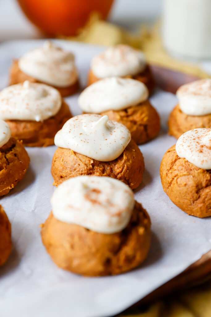 Close up of pumpkin cookies with cream cheese frosting and a dusting of cinnamon served on a parchment lined wooden plate.