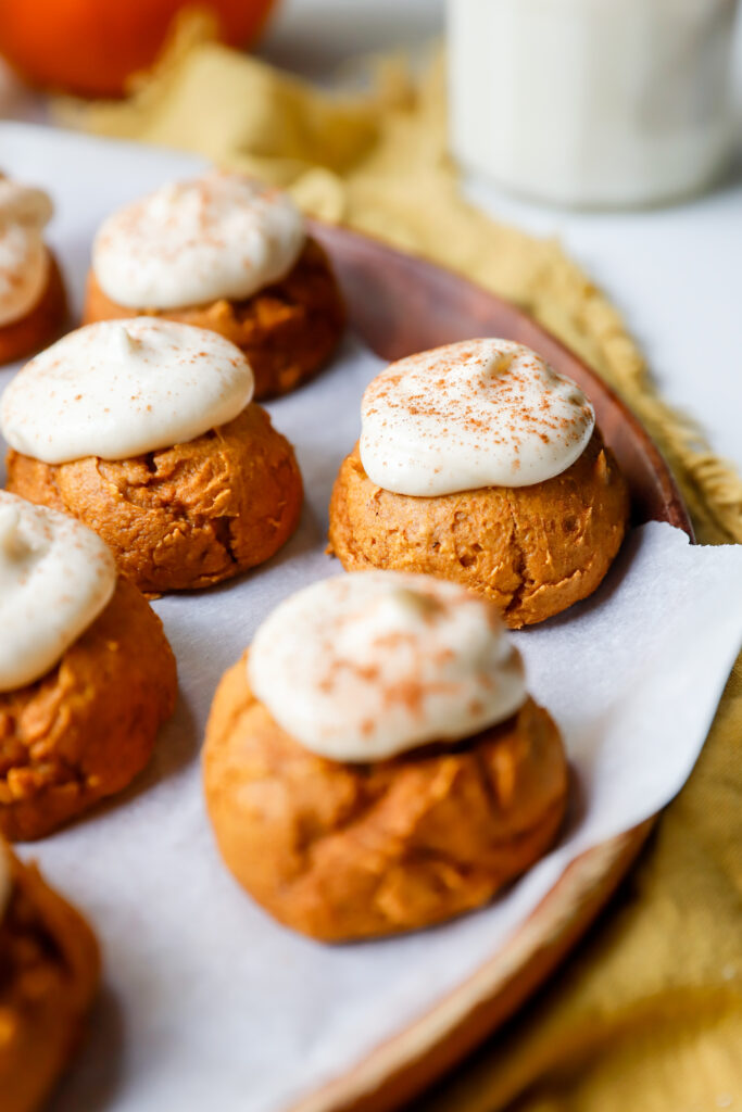Close up of pumpkin cookies with cream cheese frosting and a dusting of cinnamon served on a parchment lined wooden plate.