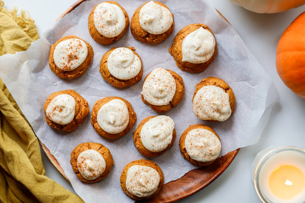 Soft pumpkin cookies with cream cheese frosting and a sprinkle of cinnamon on a parchment paper lined wooden serving plate.