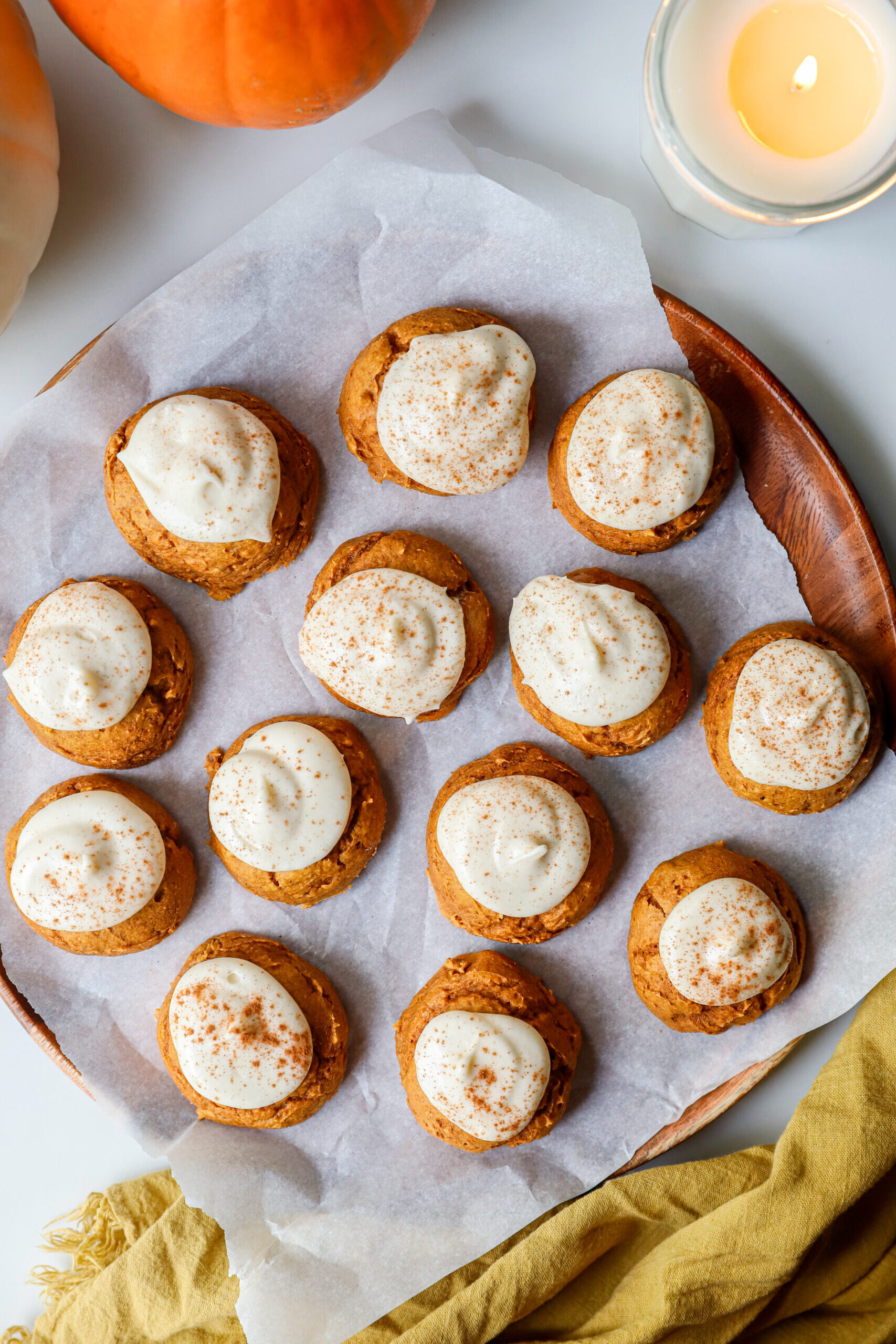 Soft pumpkin cookies with cream cheese frosting and a sprinkle of cinnamon on a parchment paper lined wooden serving plate.