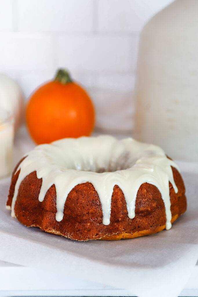 Moist pumpkin bundt cake drizzled with cream cheese icing on a parchment lined wooden platter with pumpkins behind.