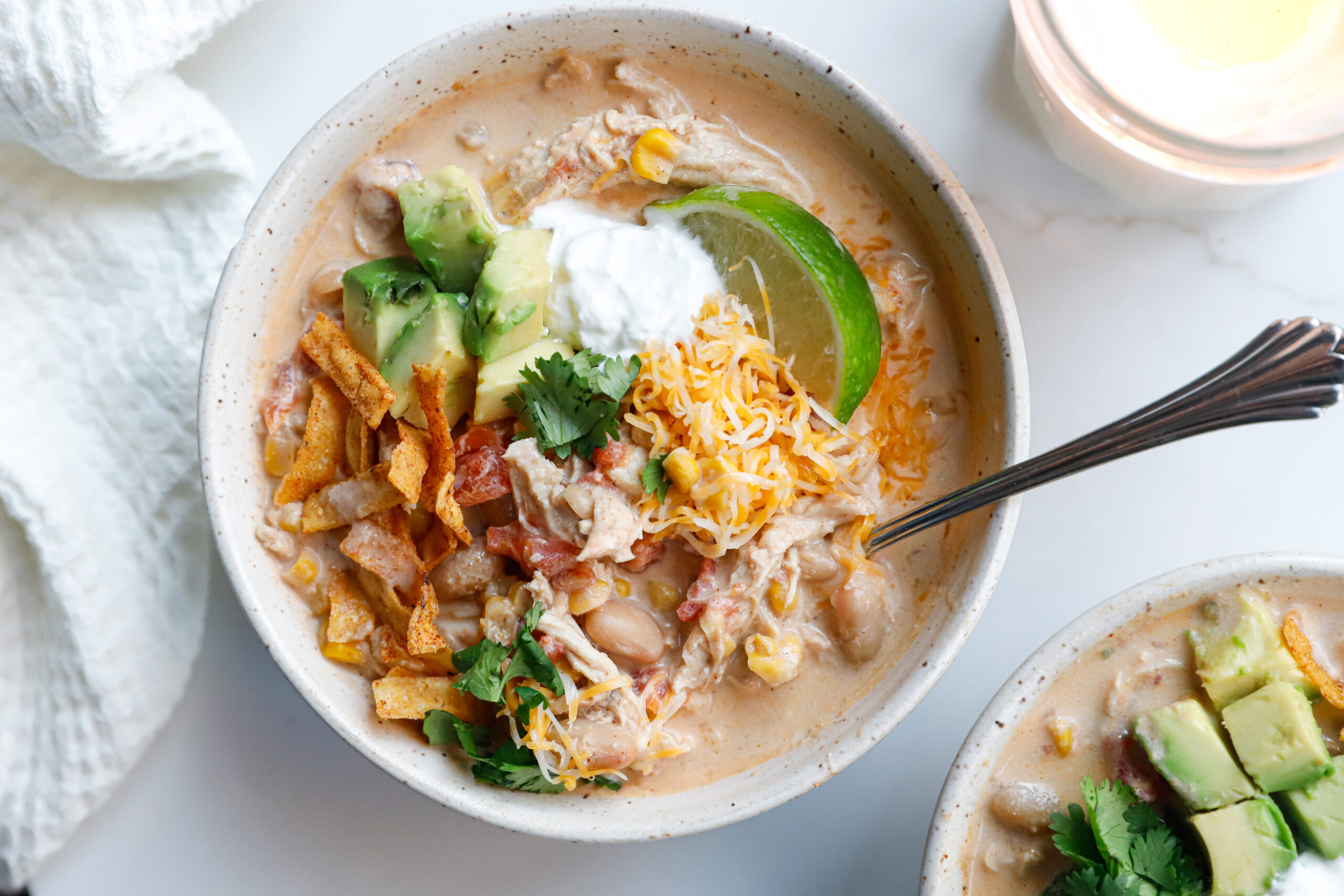 Creamy white chicken chili topped with shredded cheese, avocado, lime slice, cilantro, sour cream, and tortilla strips in a bowl, mixed sightly together with a spoon.