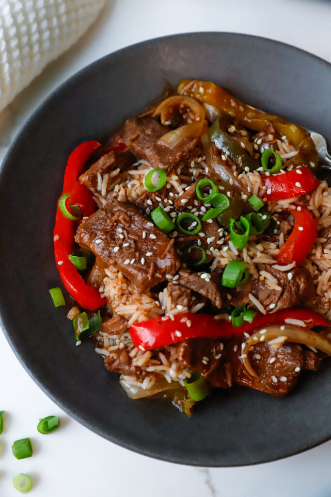 Tender slow cooker pepper steak served over rice with sesame seeds and green onions on a black plate.