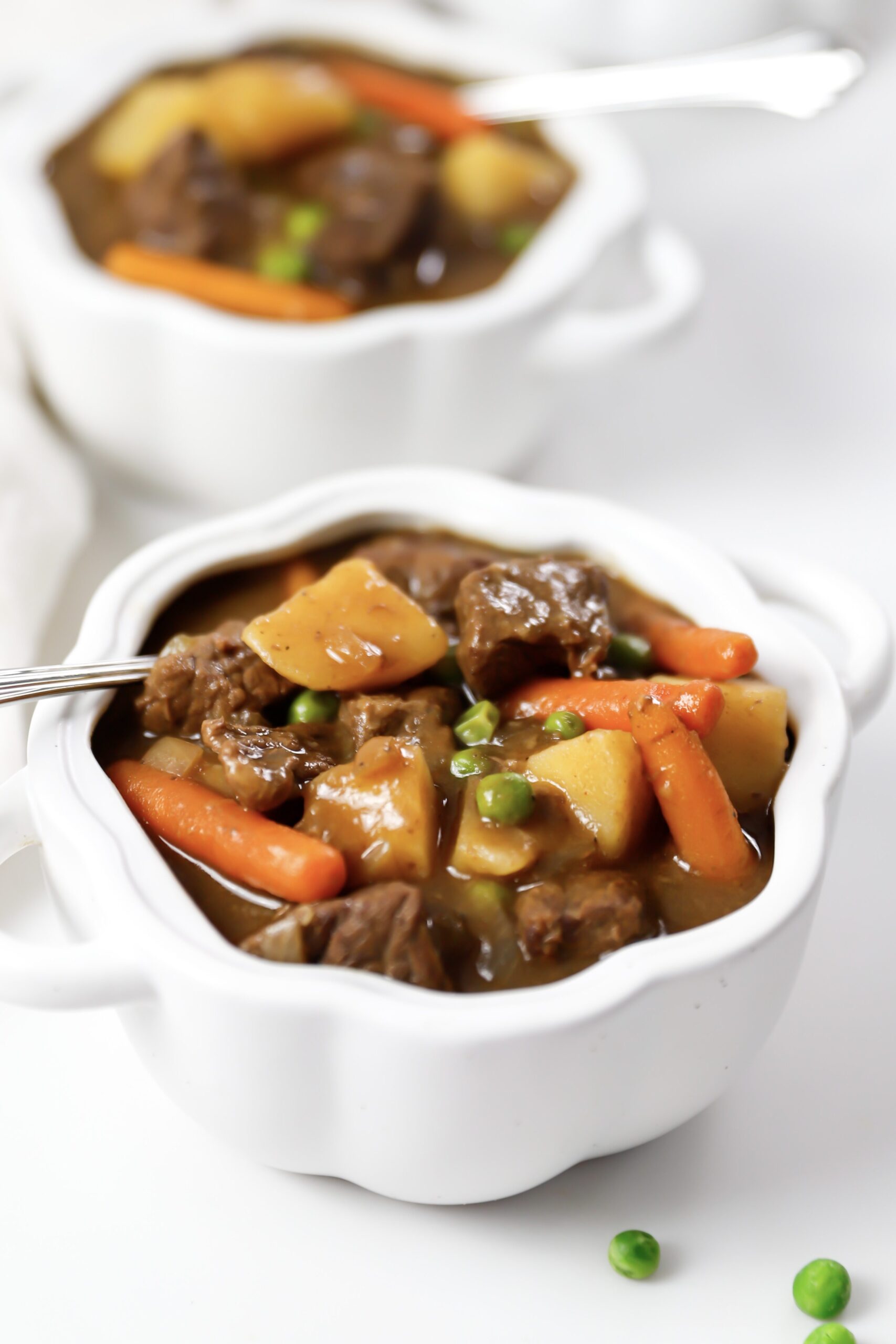 Close up shot of two bowls of slow cooker beef stew with carrots, potatoes, and peas sitting on a marble counter.