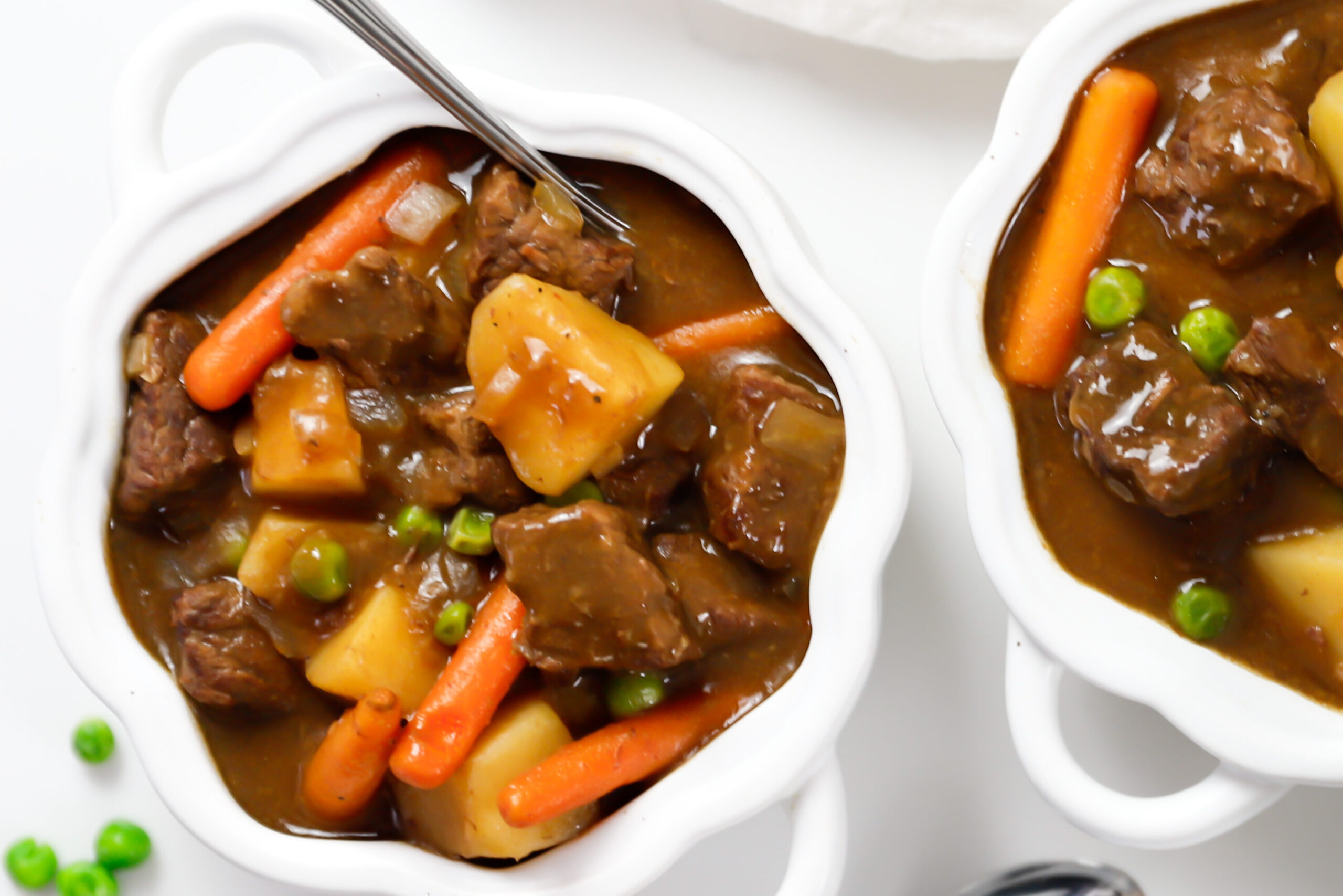 Overhead shot of two bowls of slow cooker beef stew with carrots, potatoes, and peas sitting on a marble counter.
