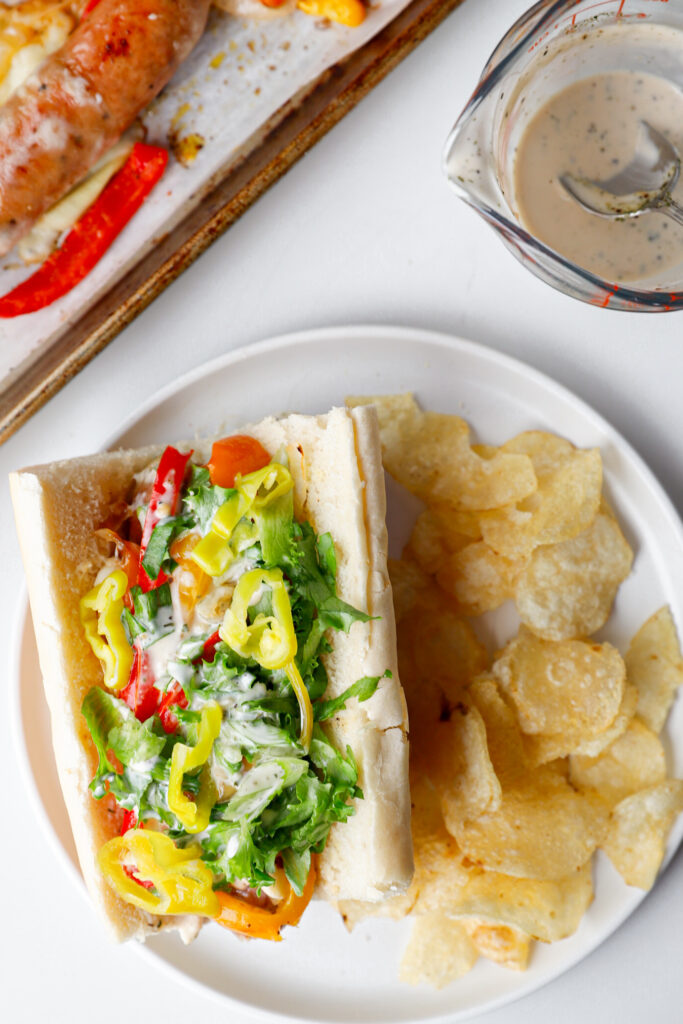 Sausage and pepper grinder sub topped with shredded lettuce and banana peppers served with a side of potato chips.