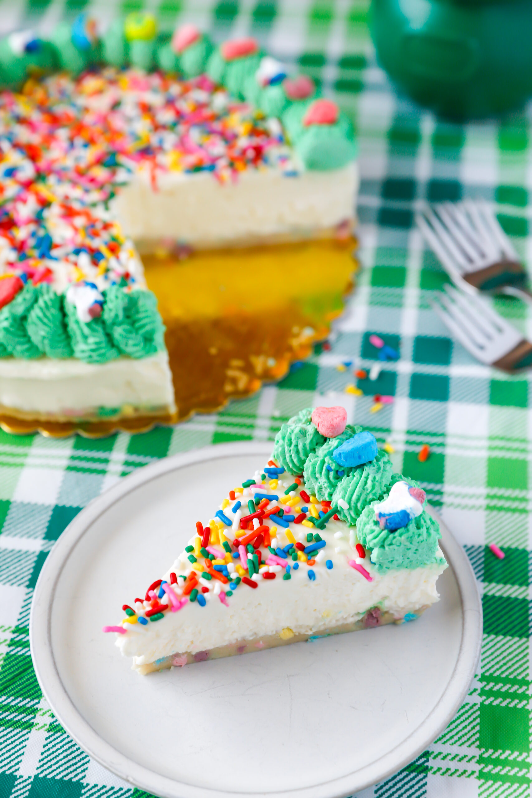 Slice of no bake Lucky Charms cheesecake with cookie dough crust, rainbow sprinkles, and whipped cream topping