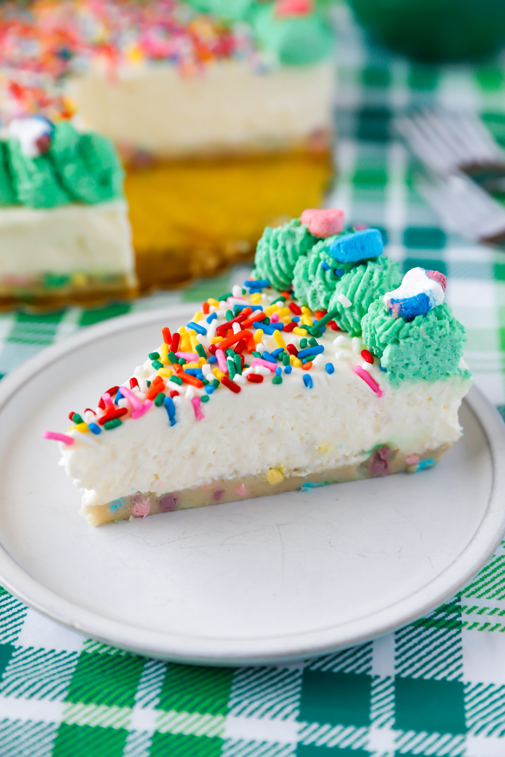 Slice of no bake Lucky Charms cheesecake with cookie dough crust, rainbow sprinkles, and whipped cream topping