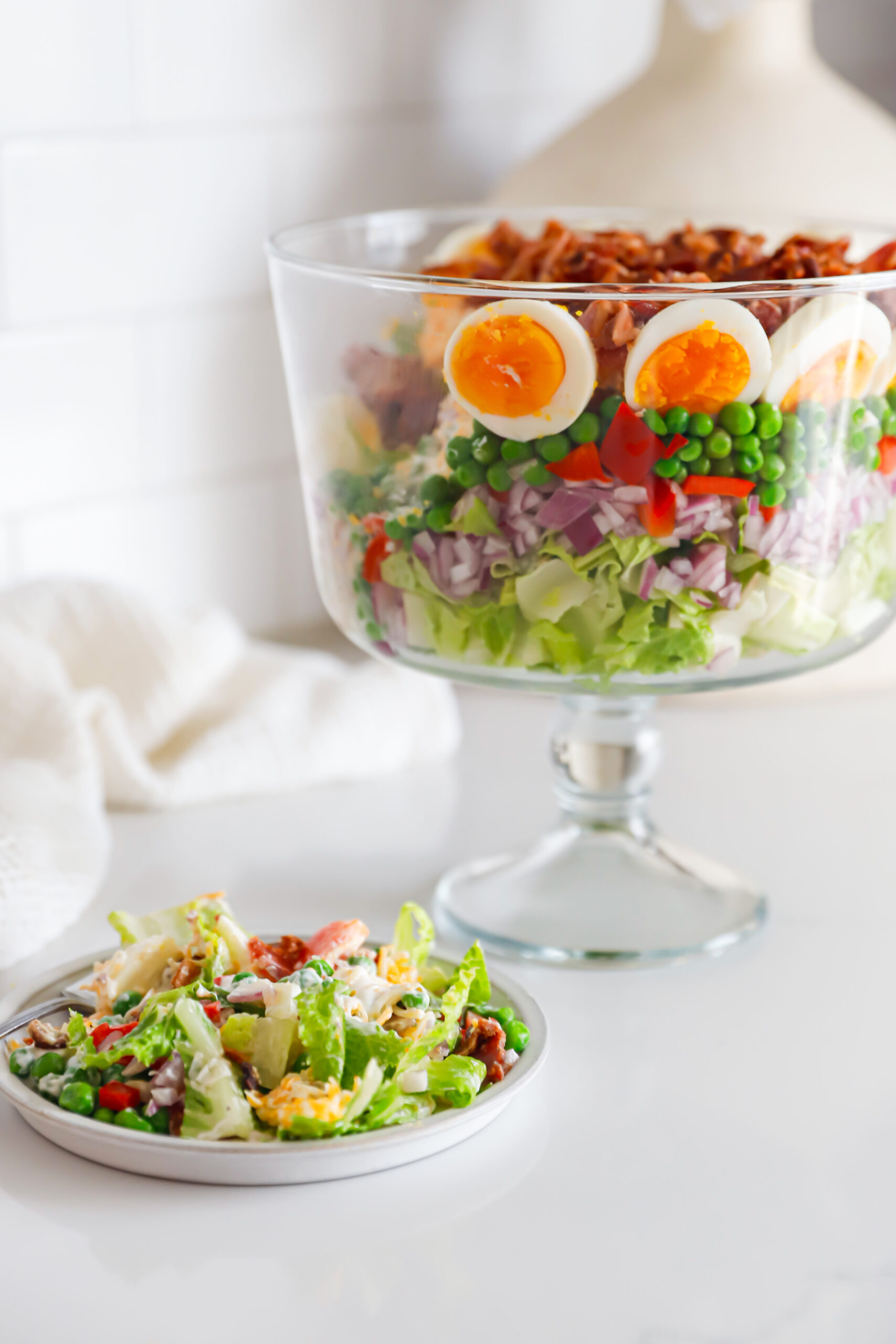 Layered salad in glass bowl with plated serving in front.